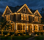 Nighttime photograph of a large two-story stone suburban house outlined in warm white holiday lights, with landscaped yard and dark blue sky.