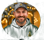 Circular-cropped photograph of a smiling man wearing a black cap and dark-green hoodie, with warm holiday-light bokeh behind him.