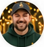 Circular portrait of a smiling bearded man in a cap and dark outerwear, against warm decorative lighting.